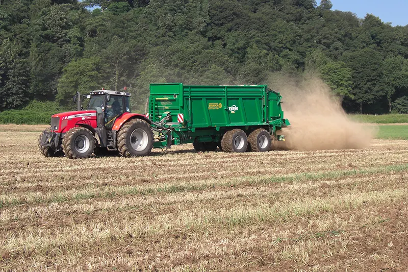 Roter Traktor mit grünem Streuer bringt Kalk auf einem Stoppelfeld aus, dahinter eine Staubwolke.