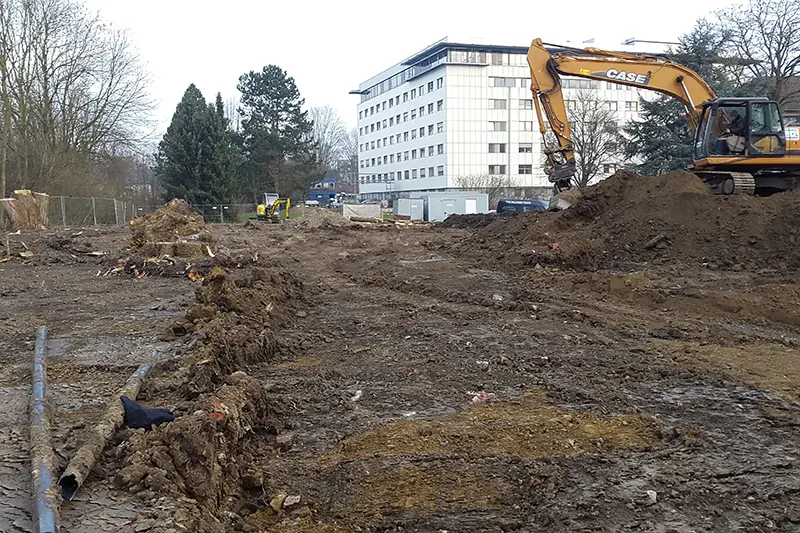 Abgeräumte Baufläche mit Erdhaufen und Wurzelresten, rechts ein Case-Kettenbagger, im Hintergrund ein mehrgeschossiges Gebäude.