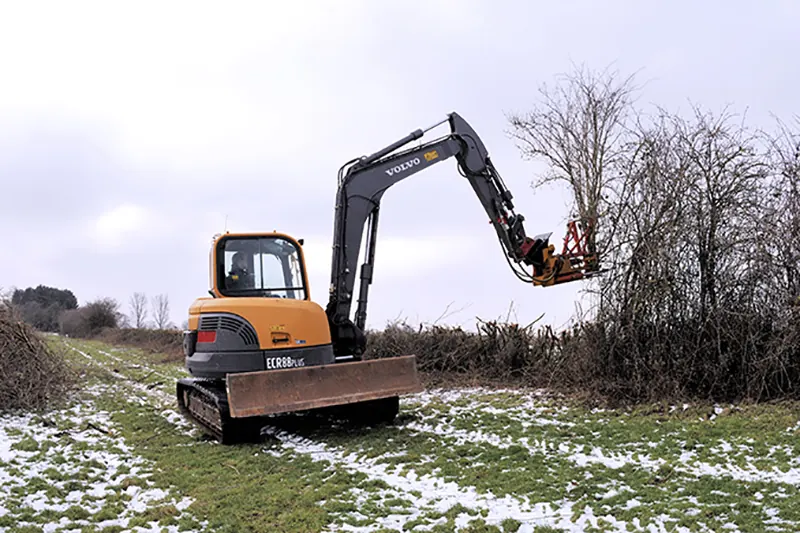 Kleiner Kettenbagger mit Anbaugerät an einer Hecke im Winter.
