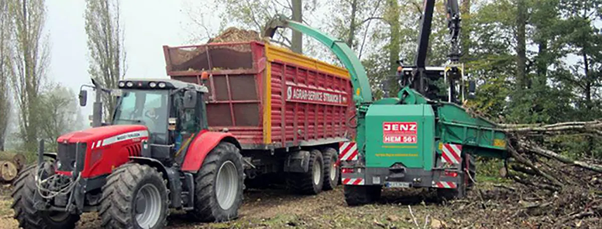 Roter Traktor mit Häckselwagen von Agrar-Service Strauch und ein grüner Jenz-Holzhacker bei der Arbeit im Wald.