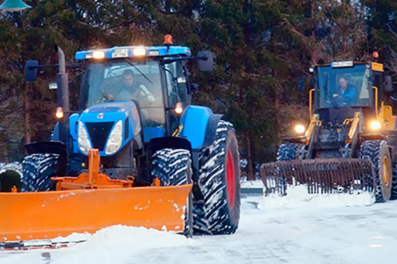 Blauer New-Holland-Traktor mit orangefarbenem Schneeschild in der Dämmerung im Schnee, rechts dahinter ein Radlader.
